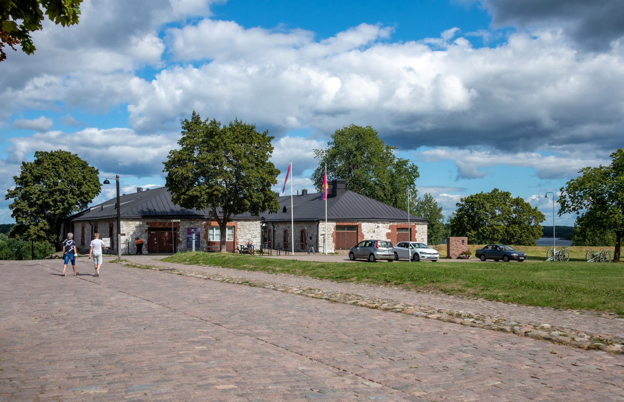 Etelä-Karjalan museo ulkoapäin kesäisessä maisemassa. Kaksi ihmistä lähestyy museota tietä pitkin.